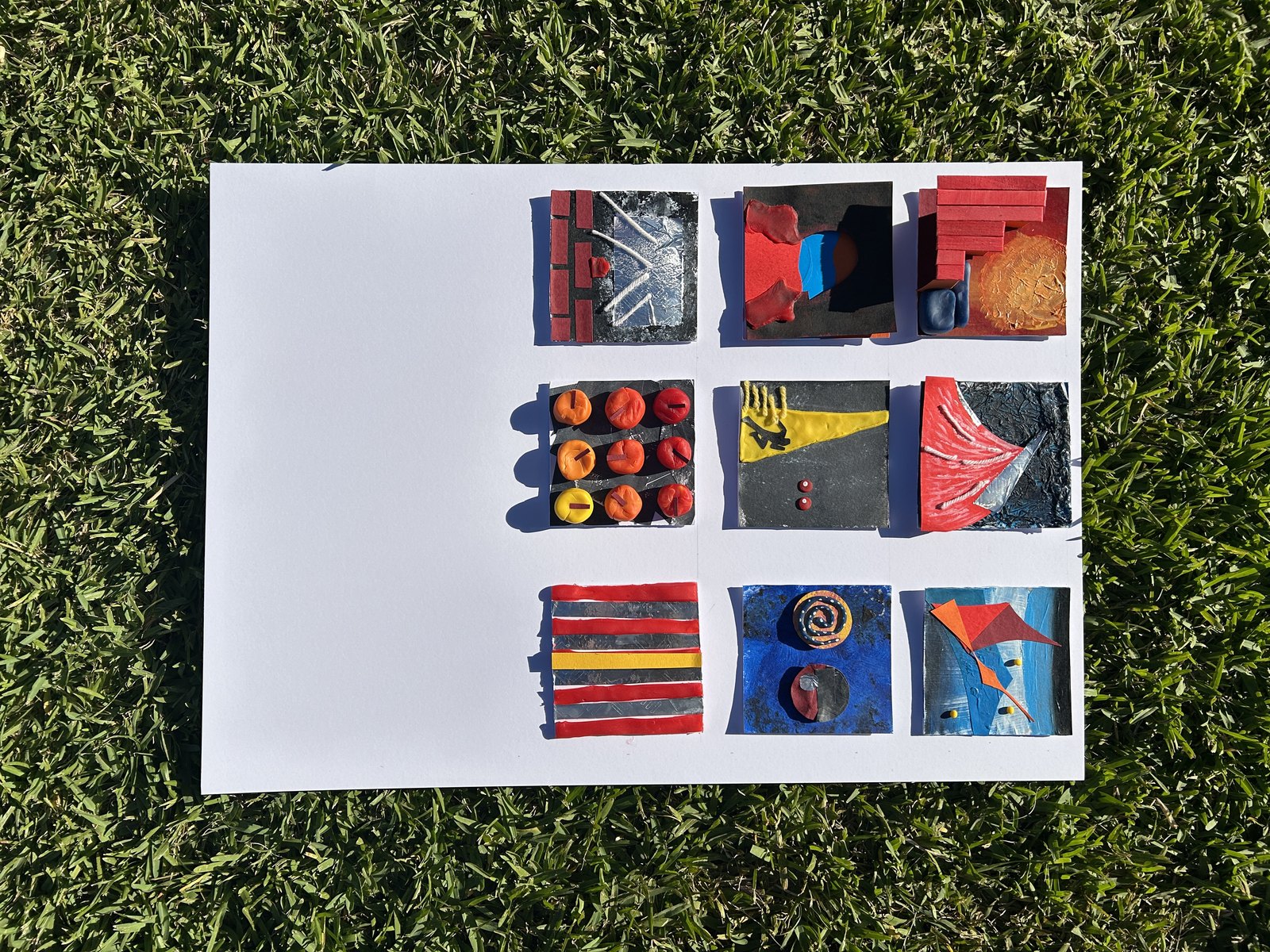 Nine mixed-media tiles laid out on white paper on grass.