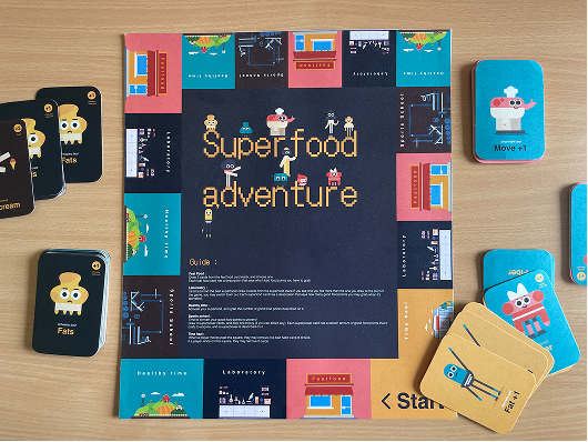 The printed Superfood Adventure board laid out on a table with its card decks and standees.